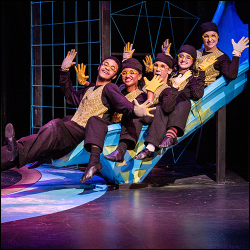 Five student actors in matching brown slacks, gold vests and gloves posing on-stage sliding down a blue slide