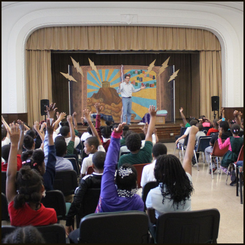 A WSU graduate acting student stands in front of a colorful on-stage backdrop while engaging with a classroom full of young students with arms raised to ask questions.